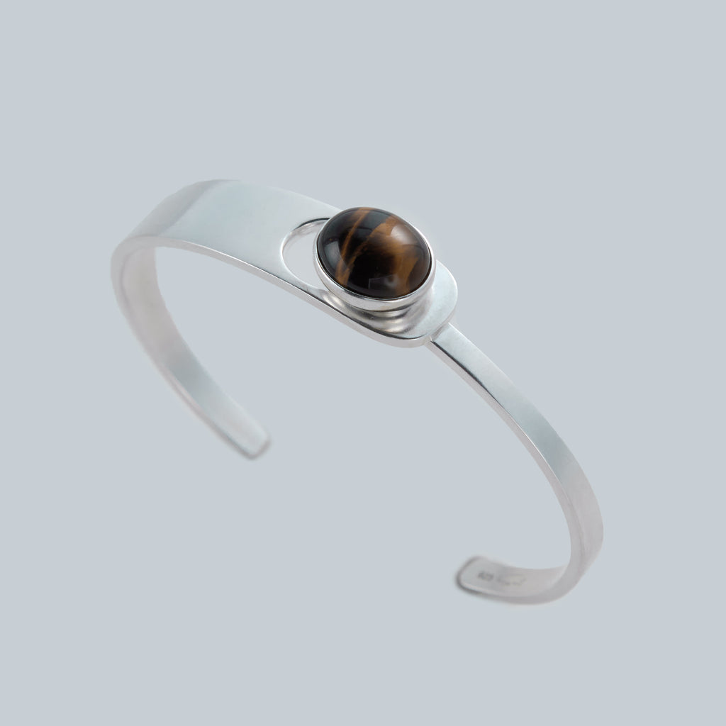 Saël Aperture Cuff in muted silver, 49*59mm interior diameter, with an elliptical cutout and bezel-set Tiger Eye