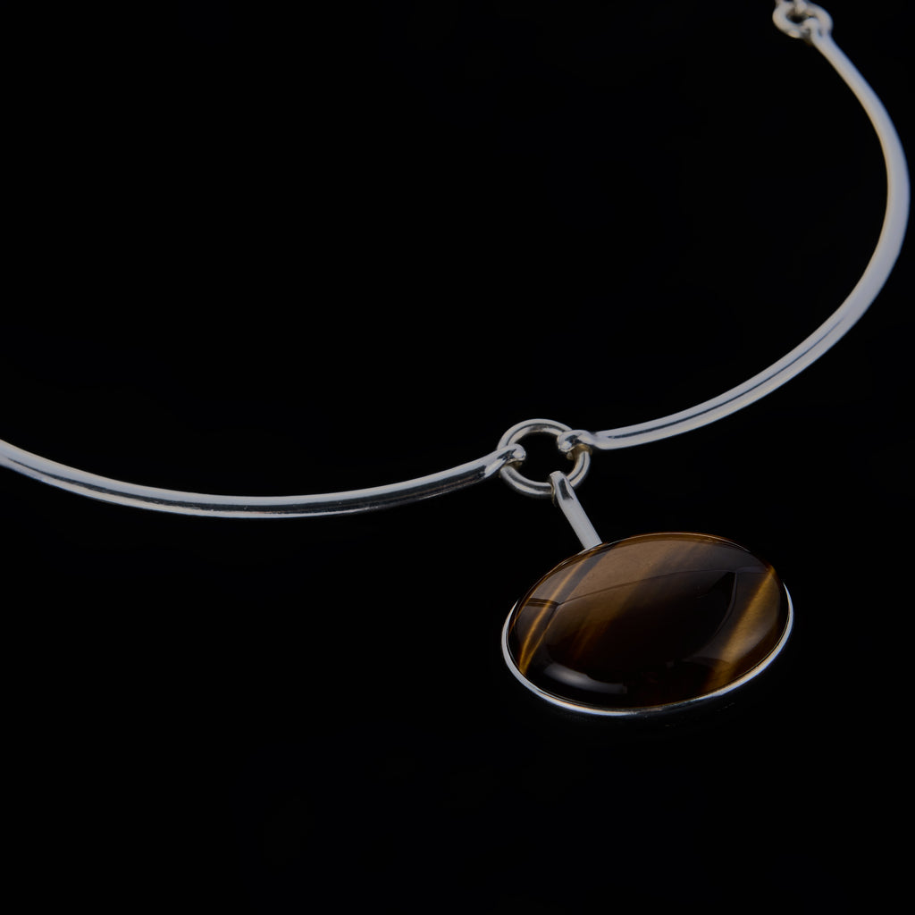 A modernist 925 silver necklace featuring a rigid frontal arc, hidden articulated chain, and a suspended Tiger Eye cabochon. Architectural jewelry by Saël.